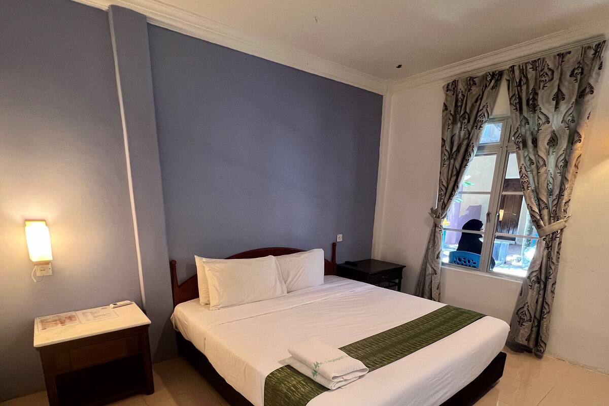 A neatly made bed with white linens and a green accent, a bedside table with a lamp, and a window with patterned curtains.