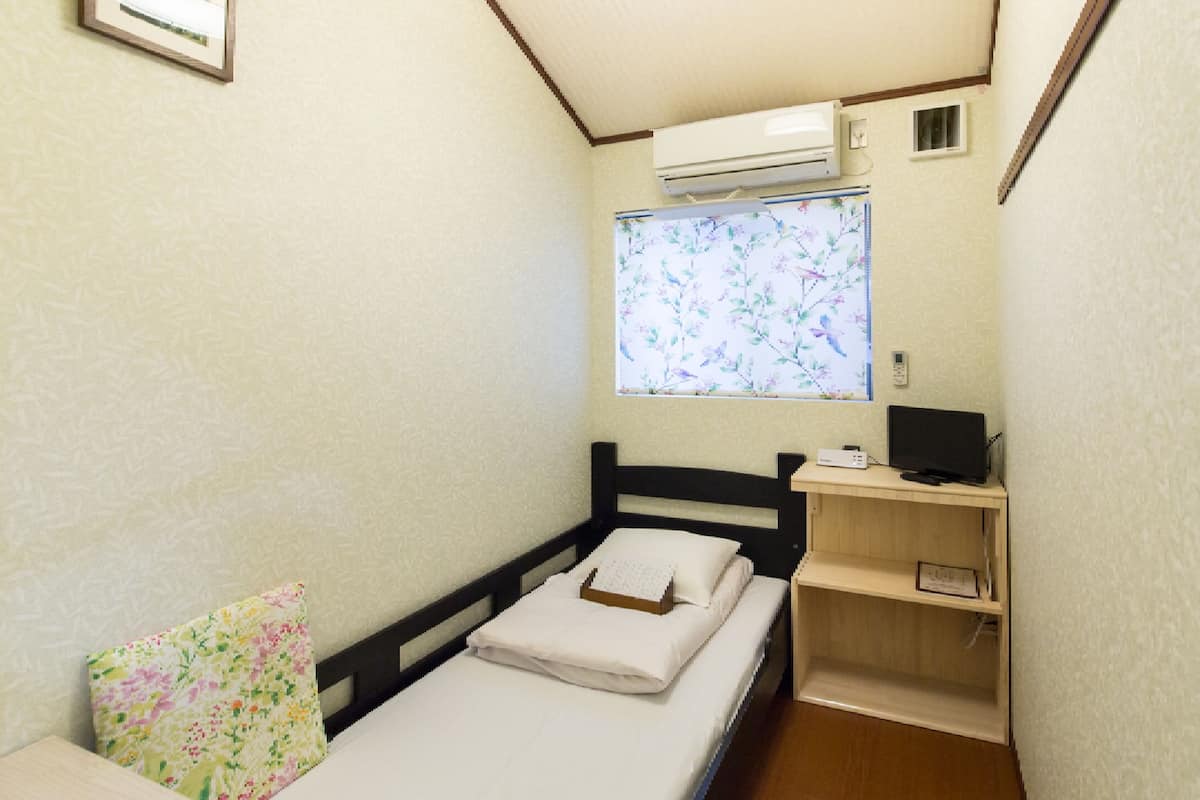 A small, narrow room with a single bed, a window with floral blinds, a desk with a computer, and a wall-mounted air conditioner.