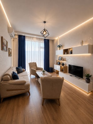 A modern living room with a sofa, chairs, a coffee table, a TV, and a built-in shelf with decorative items. - Vazi Aparthotel (Tbilisi)