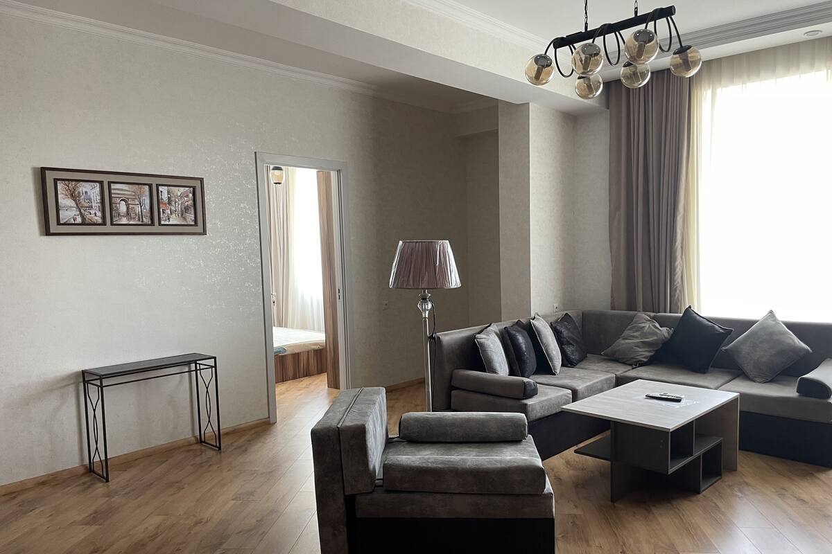 En moderne stue med sofa, skammel og sofabord. Der er en lampe, et billede i ramme på væggen og en dør, der fører til et andet rum.