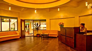 A reception area with a wooden reception desk, a sofa, and a table. There is a plant and a vase with flowers on the desk. - The Palm Hotel (Nainital)