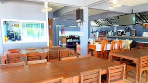 A restaurant with wooden tables and chairs, a kitchen area, and a large window with a view. - Tharadol Beach Resort (Tha Sala)