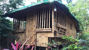 Una cabaña rústica de bambú con techo de paja, rodeada de abundante follaje y plantas en macetas.