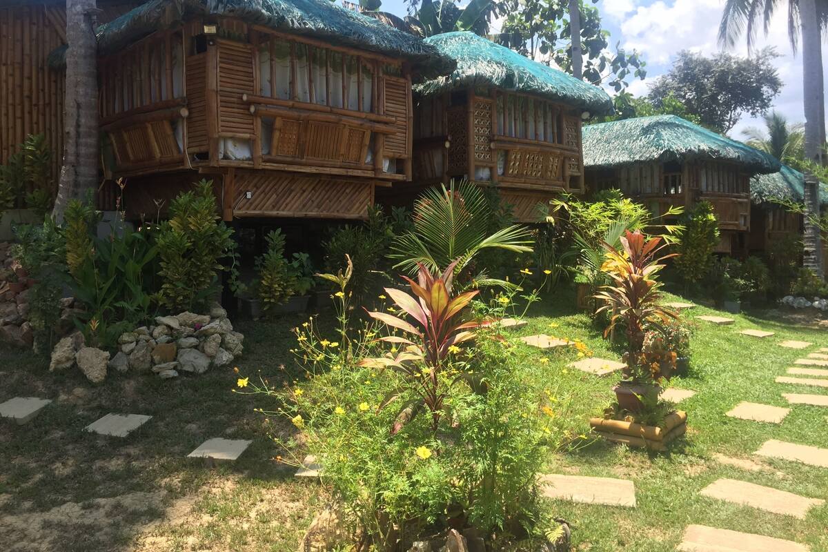 Resort com edifícios de madeira, telhados de palha e um jardim com plantas tropicais.