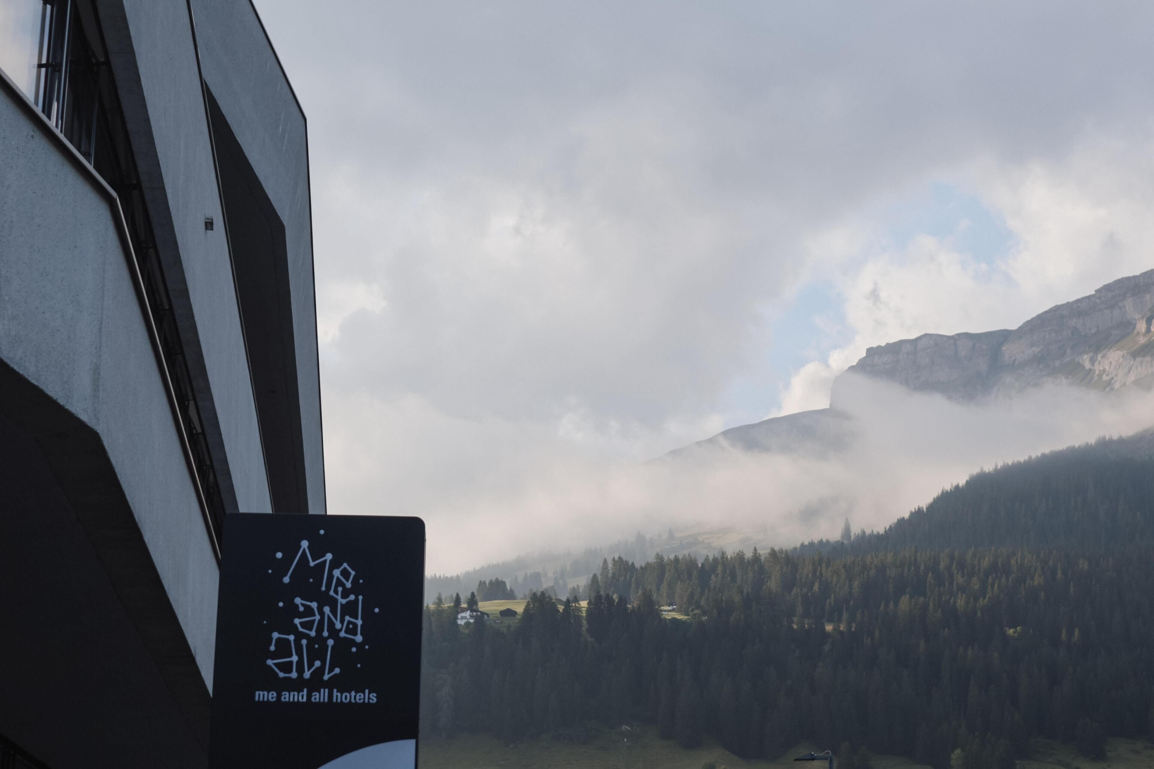 A mountainous landscape with a forested valley and a building with a sign reading "me and all hotels".