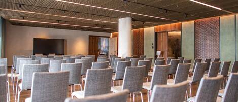 A conference room with rows of grey chairs, a large pillar, and a flat-screen TV on the wall.