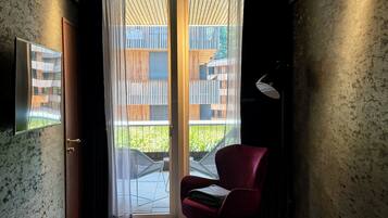 Une pièce compacte avec une porte-fenêtre coulissante donnant sur un espace extérieur, une chaise rouge, une petite table avec un magazine et un lit recouvert d’une couverture verte.