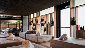 A modern spa room with multiple massage beds, wooden shelves with plants, and a large window providing natural light.
