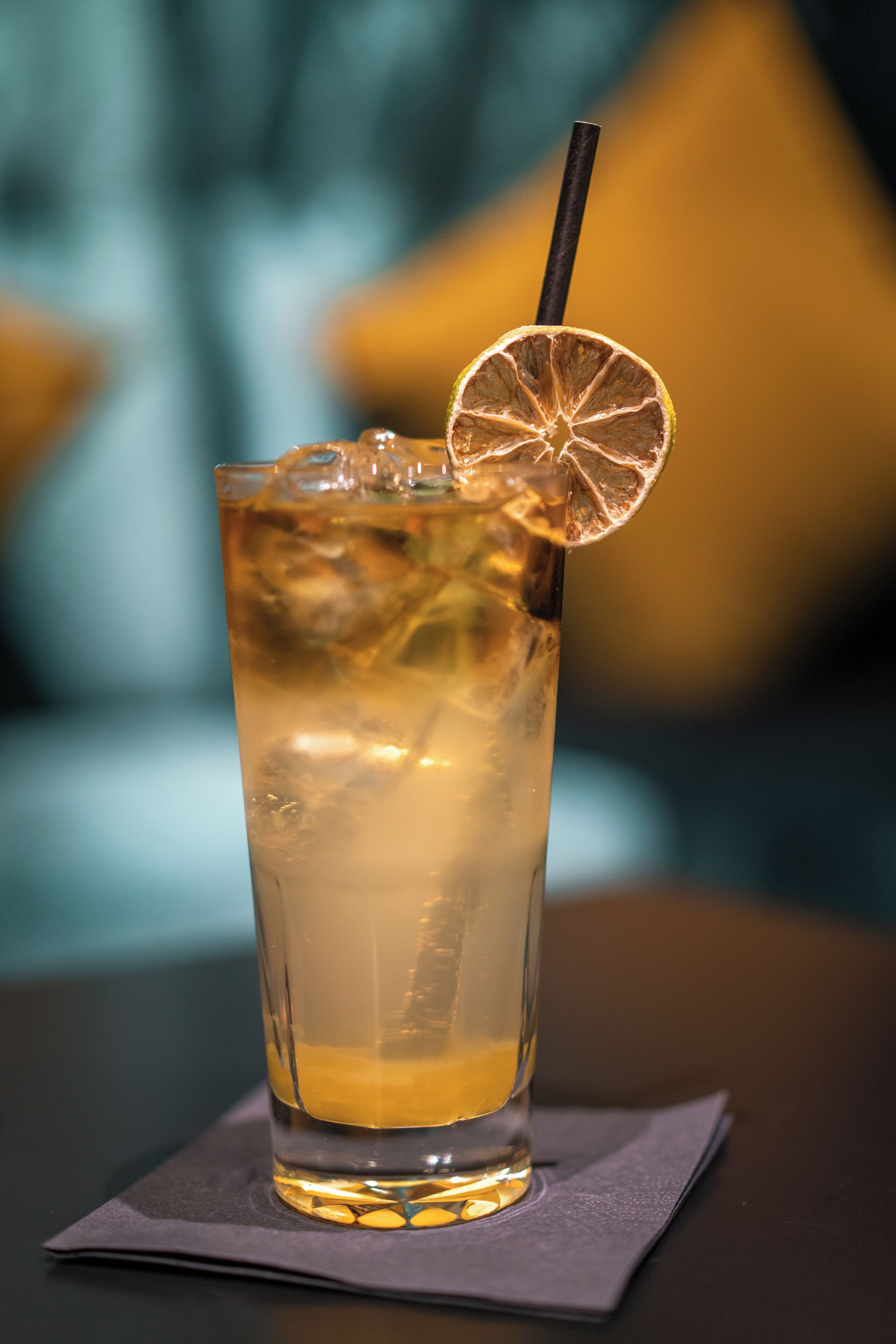 A tall glass of iced beverage with a lemon slice and straw on a napkin.