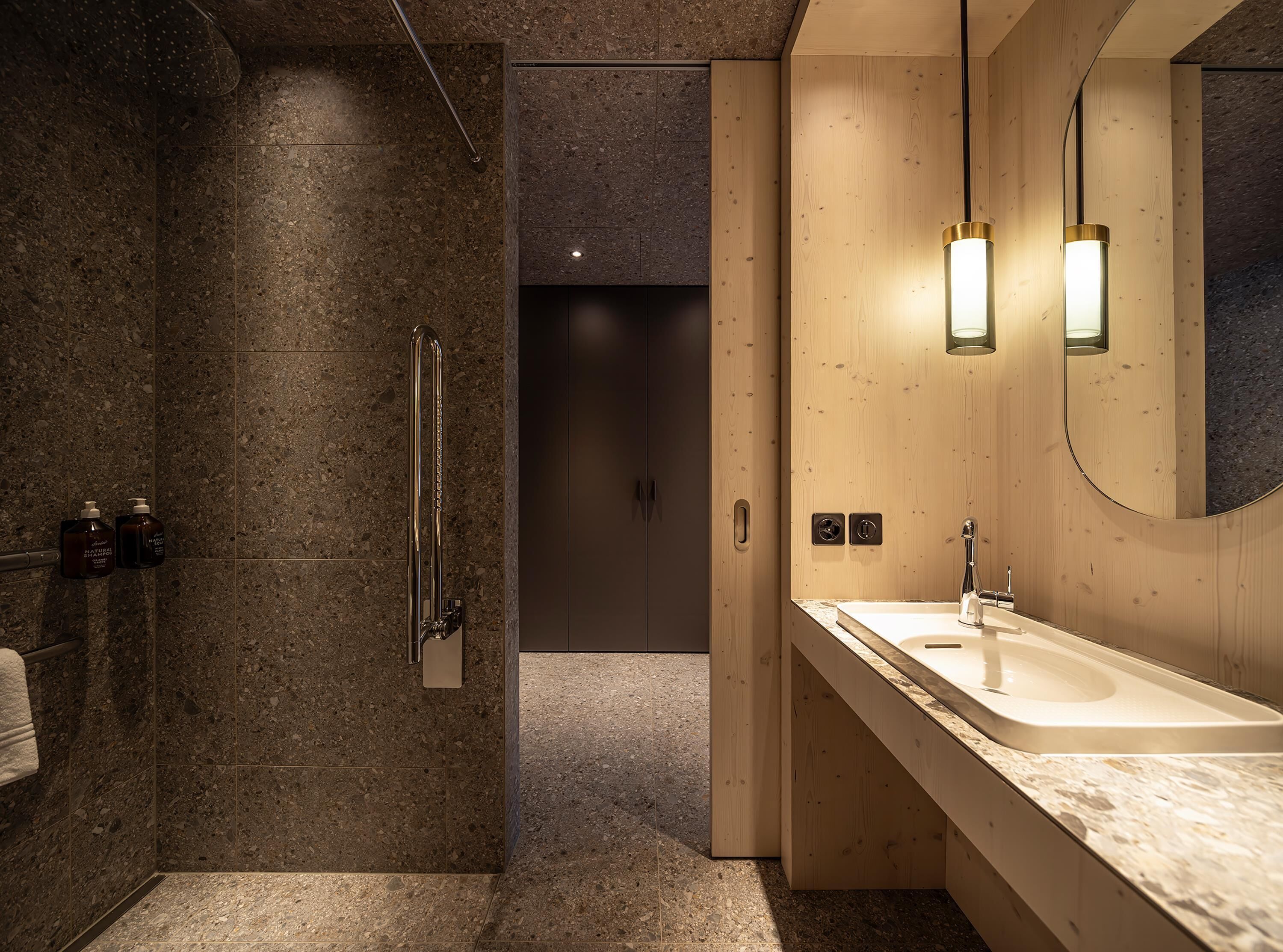 A modern bathroom with a granite shower, a wooden vanity with a sink, and a mirror with wall-mounted lights.