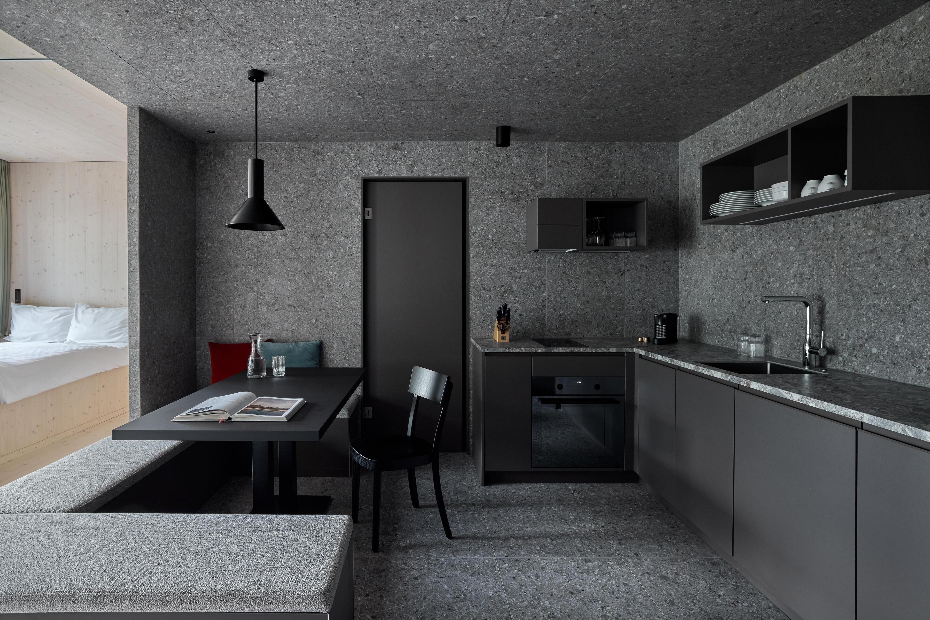 A modern kitchen with a built-in oven, a sink, and a countertop. There is a dining area with a bench and a table, and a chair. The kitchen has a dark color scheme with gray countertops and cabinets.
