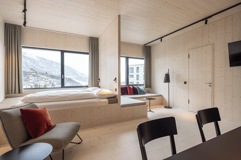 A modern room with a large window, a bed, a seating area, and a TV.