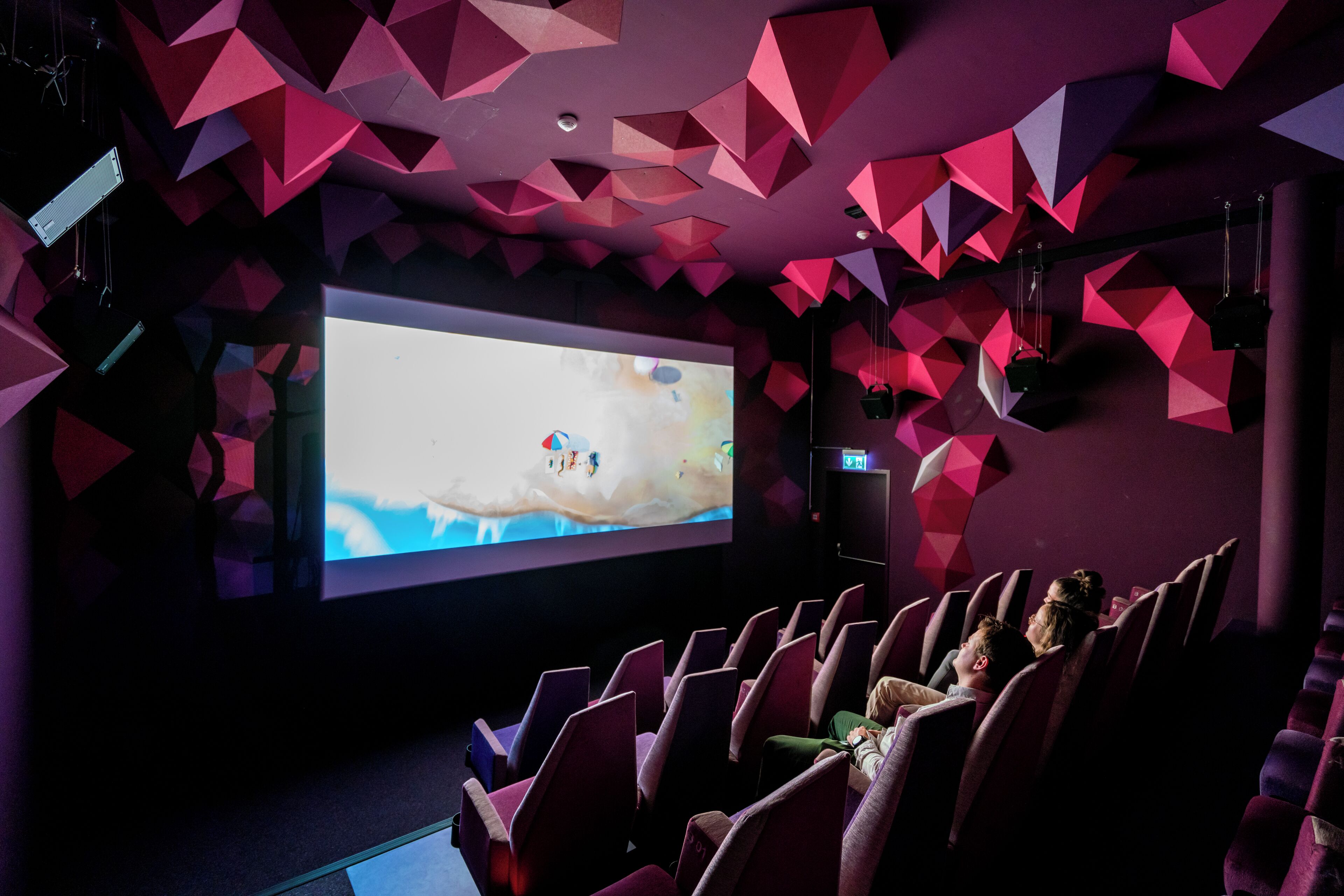 Une salle de cinéma intérieure dotée d’un grand écran, de rangées de sièges et de décorations murales aux motifs géométriques.