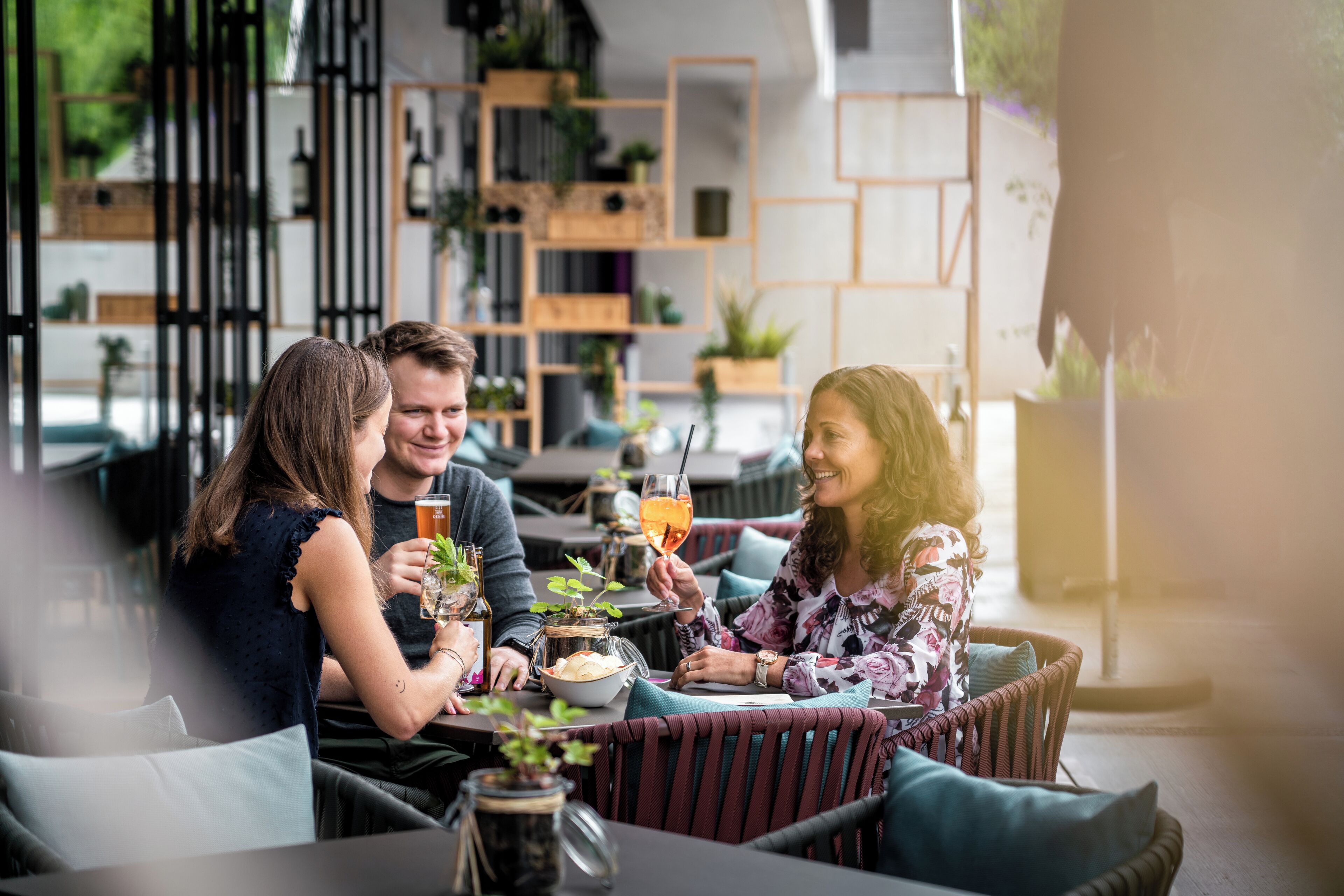 A group of people dining at an outdoor cafe with wicker furniture and shelves with plants in the background.