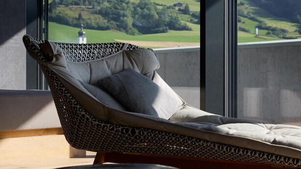 A wicker lounge chair with cushions, positioned near a large window overlooking a scenic landscape with mountains and a golf course.