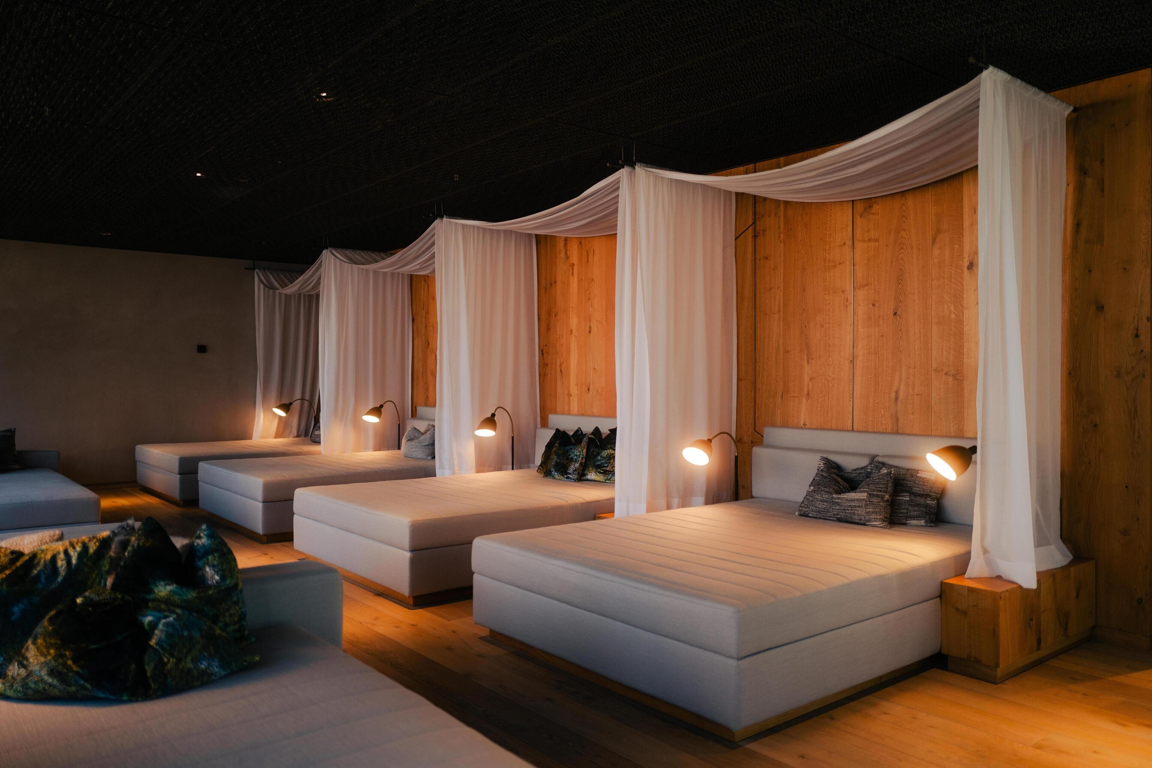 A room with three beds, each with white bedding and draped white curtains. There are bedside lamps and a wooden headboard on the right side.