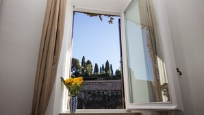 A window with beige curtains, a vase with yellow flowers, and a clear view of trees and a building outside. - Fori Romani B&B (Rome)
