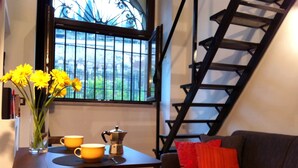 A cozy living room with a dark brown sofa, a coffee table with a vase of yellow flowers, two yellow cups, and a silver coffee pot. A black spiral staircase is visible to the right. - Fori Romani B&B (Rome)