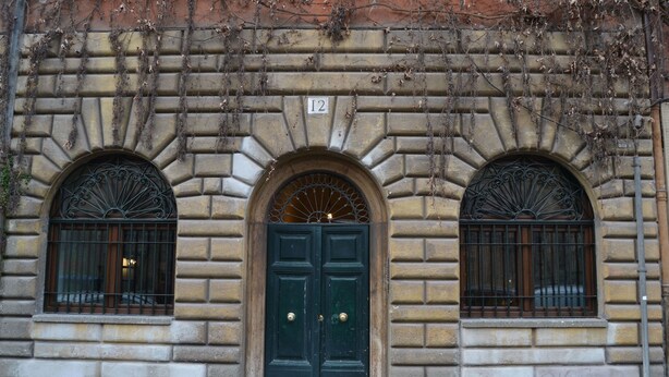 A building facade with a green double door, two arched windows with metal grilles, and a number '12' above the door. - Fori Romani B&B (Rome)