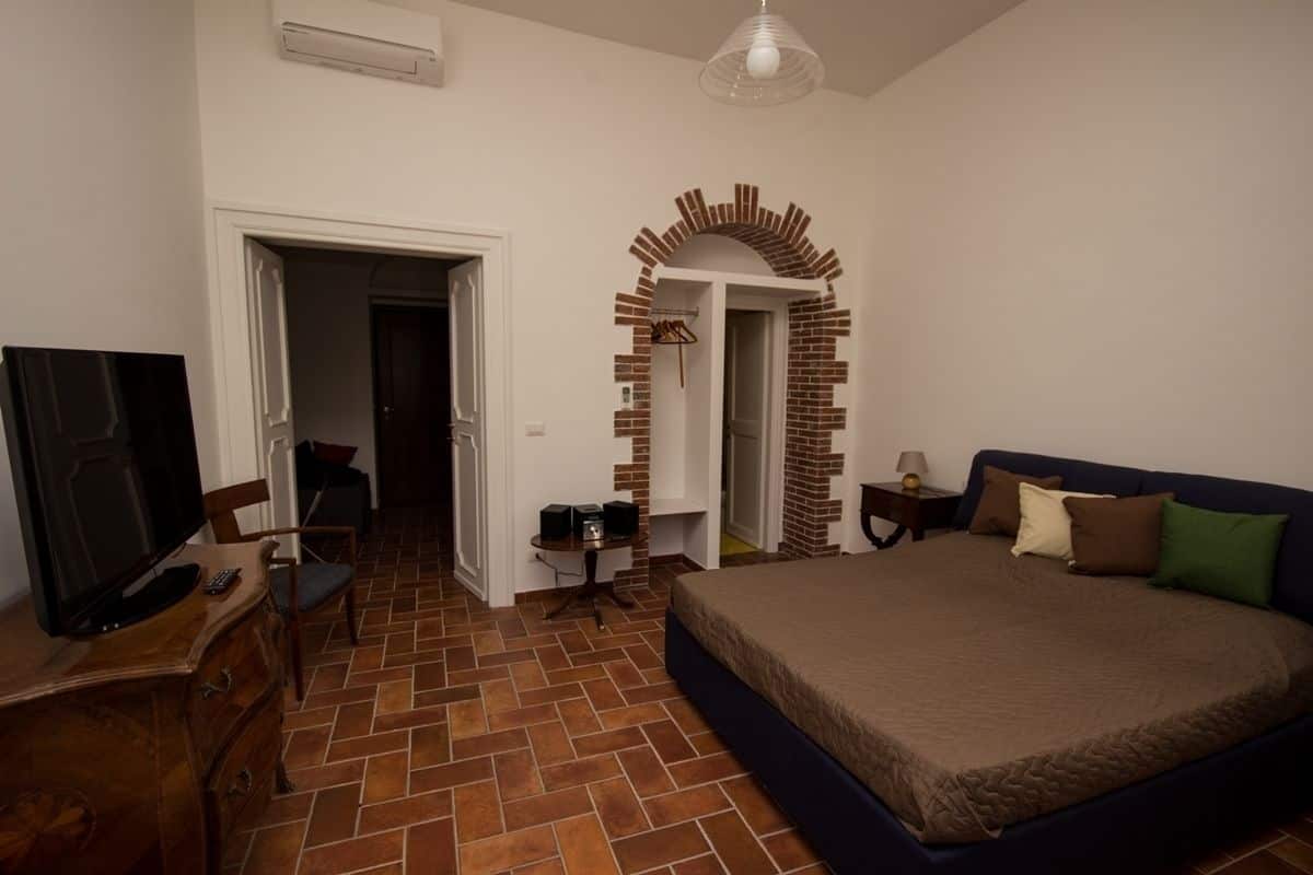 A bedroom with a large bed, a TV on a wooden dresser, a desk with a chair, and a brick archway.
