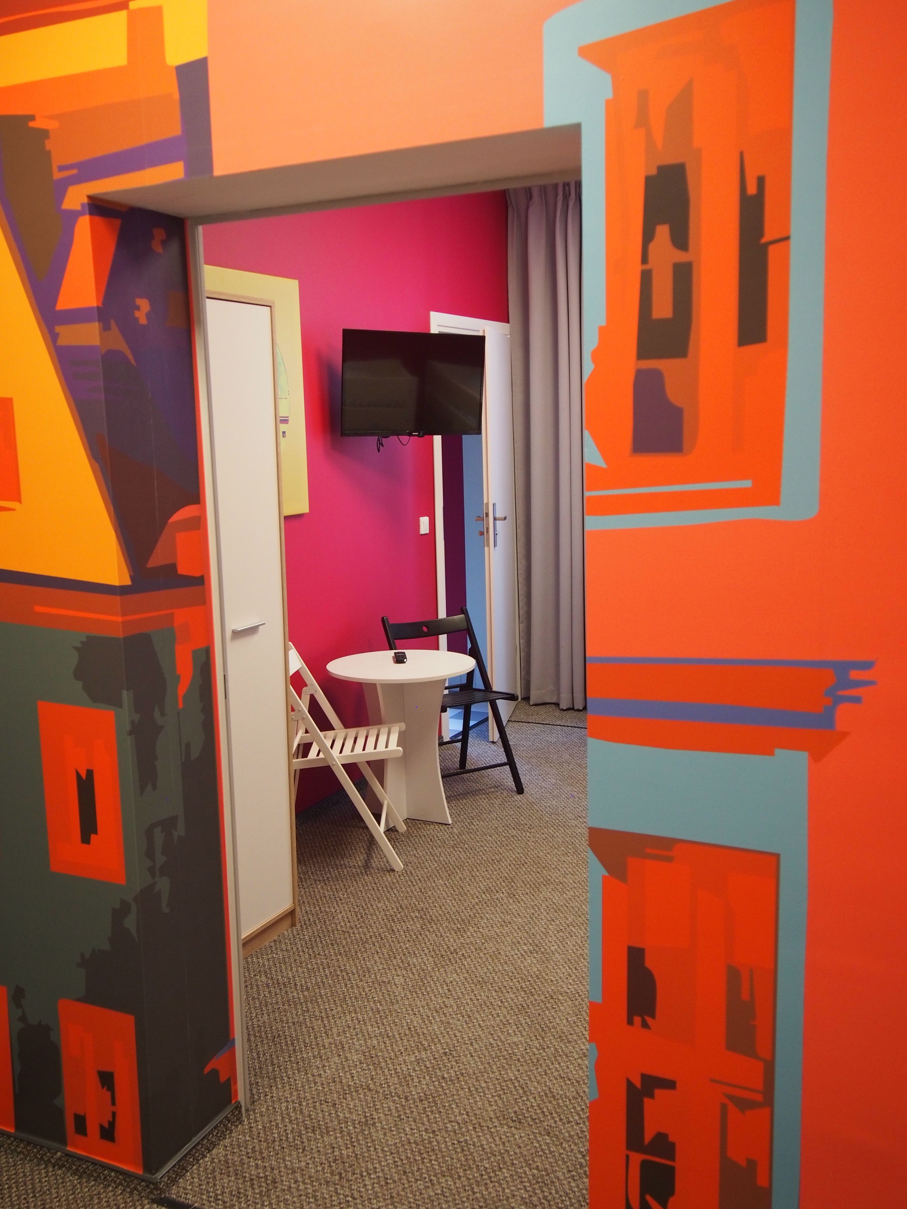 A hotel room with a vibrant color scheme, a flat-screen TV mounted on the wall, a small round table with two chairs, and a door leading to another room.