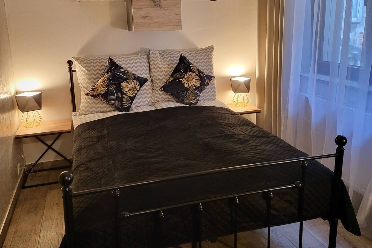 Ein Schlafzimmer mit einem Bett, Nachttischlampen, einem Wandregal mit Pflanzen und einem Fenster mit Vorhängen.