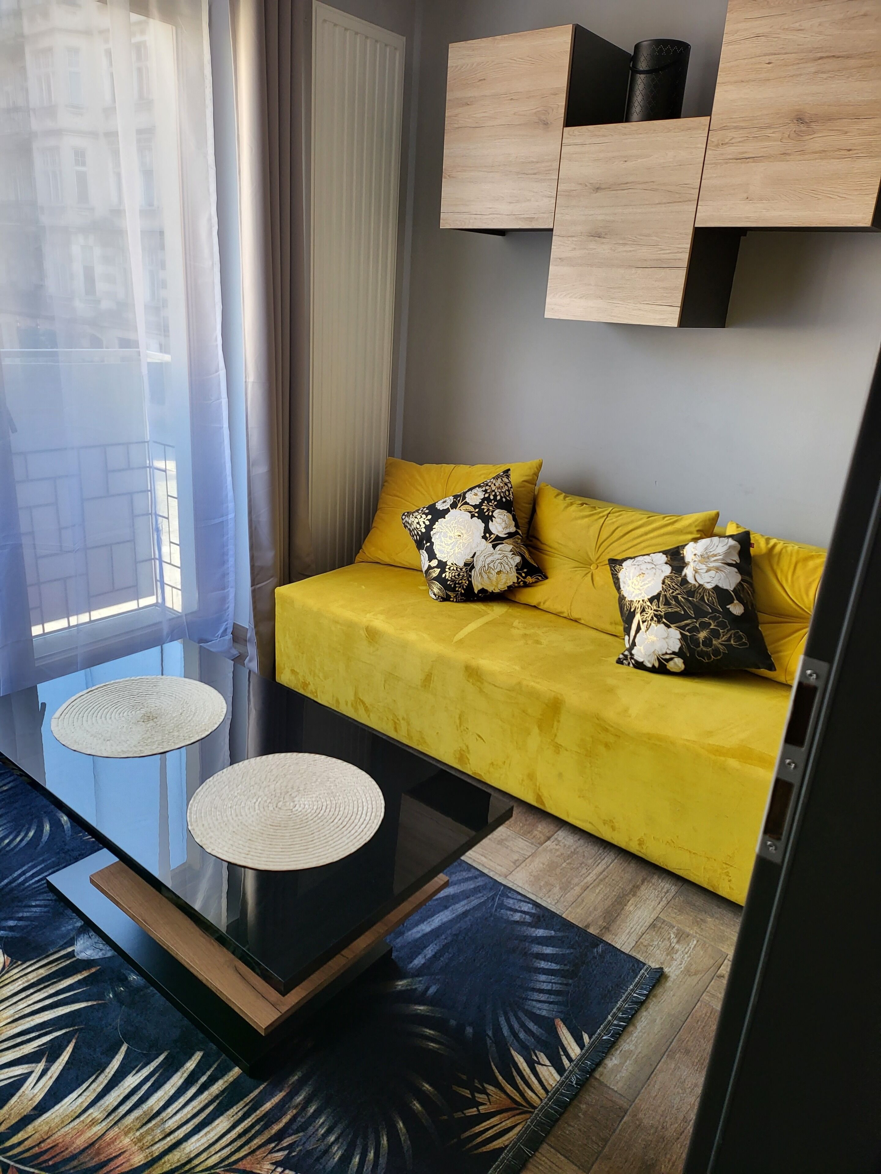 Un salon moderne avec un canapé jaune, deux coussins assortis, une table basse en verre et un tapis bleu à motifs.