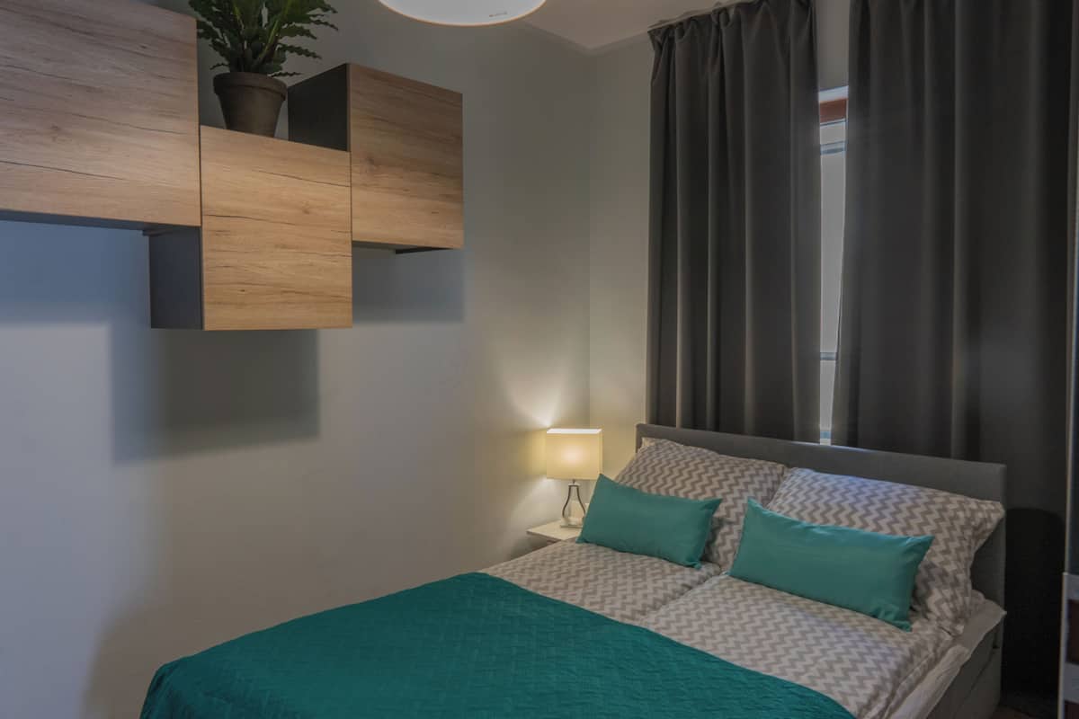 Ein Schlafzimmer mit einem Bett, einem Nachttisch, einer Wandablage mit einer Pflanze und einem Fenster mit Vorhängen.