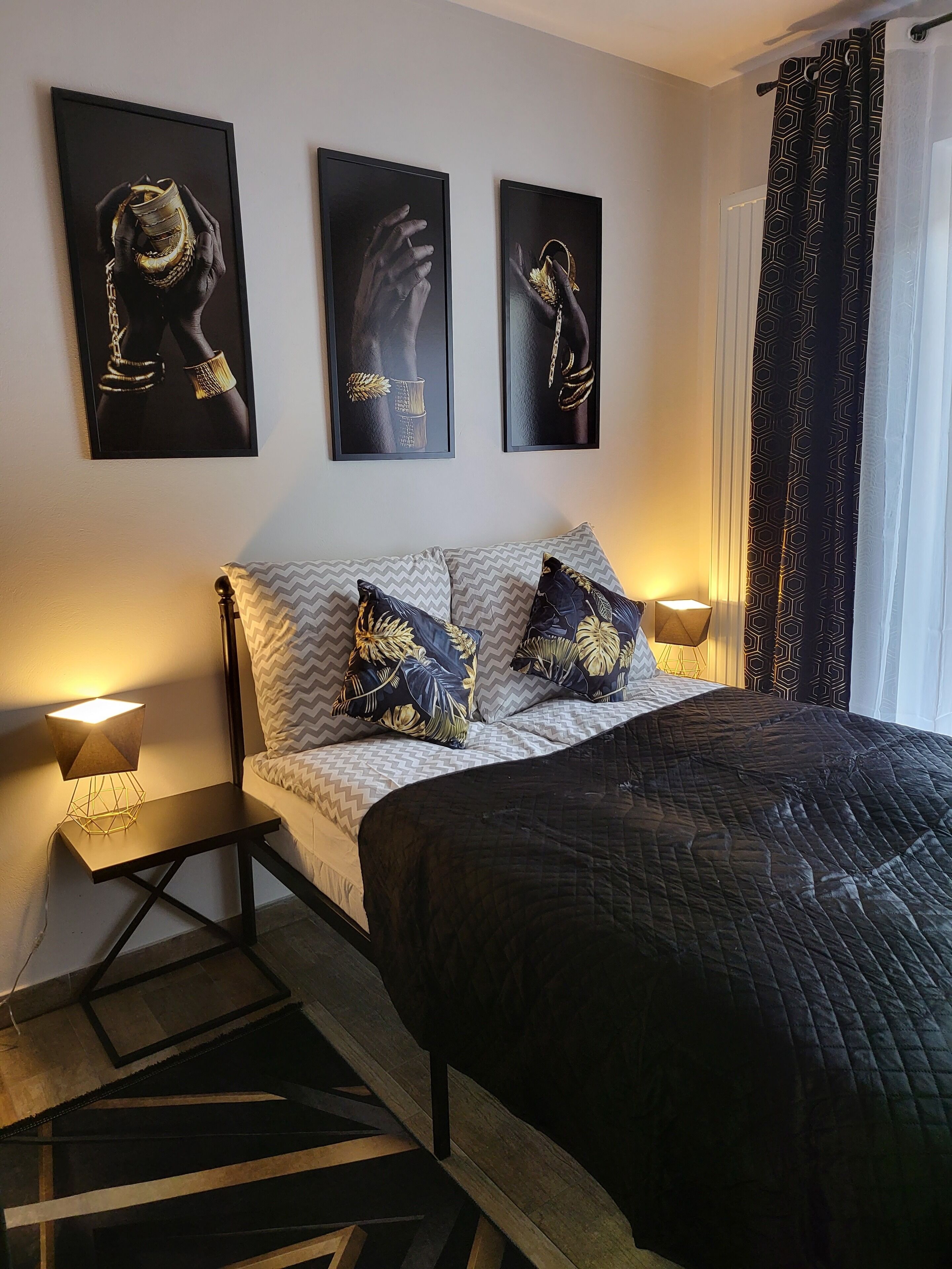 A bedroom with a bed, two wall-mounted picture frames, a patterned curtain, and a bedside lamp.