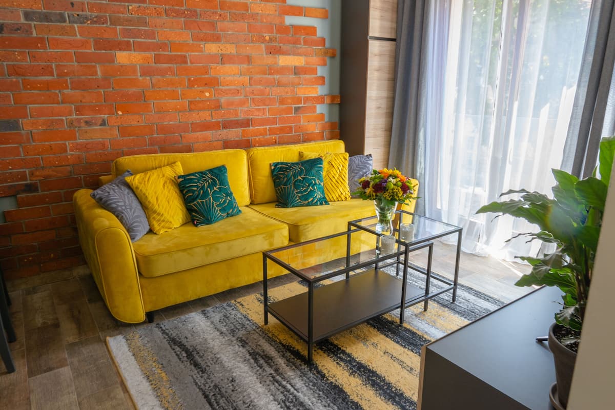 Ein gelbes Sofa mit gemusterten Kissen, ein Glastisch und ein Blumentopf.
