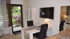 A hotel room with a dining table, chairs, a TV mounted on the wall, a bed with a headboard, and a view of a garden with flowers. - Ferienwohnung Schelsen (Moenchengladbach)