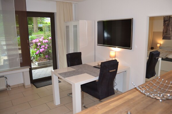 A hotel room with a dining table, chairs, a TV mounted on the wall, a bed with a headboard, and a view of a garden with flowers. - Ferienwohnung Schelsen (Moenchengladbach)