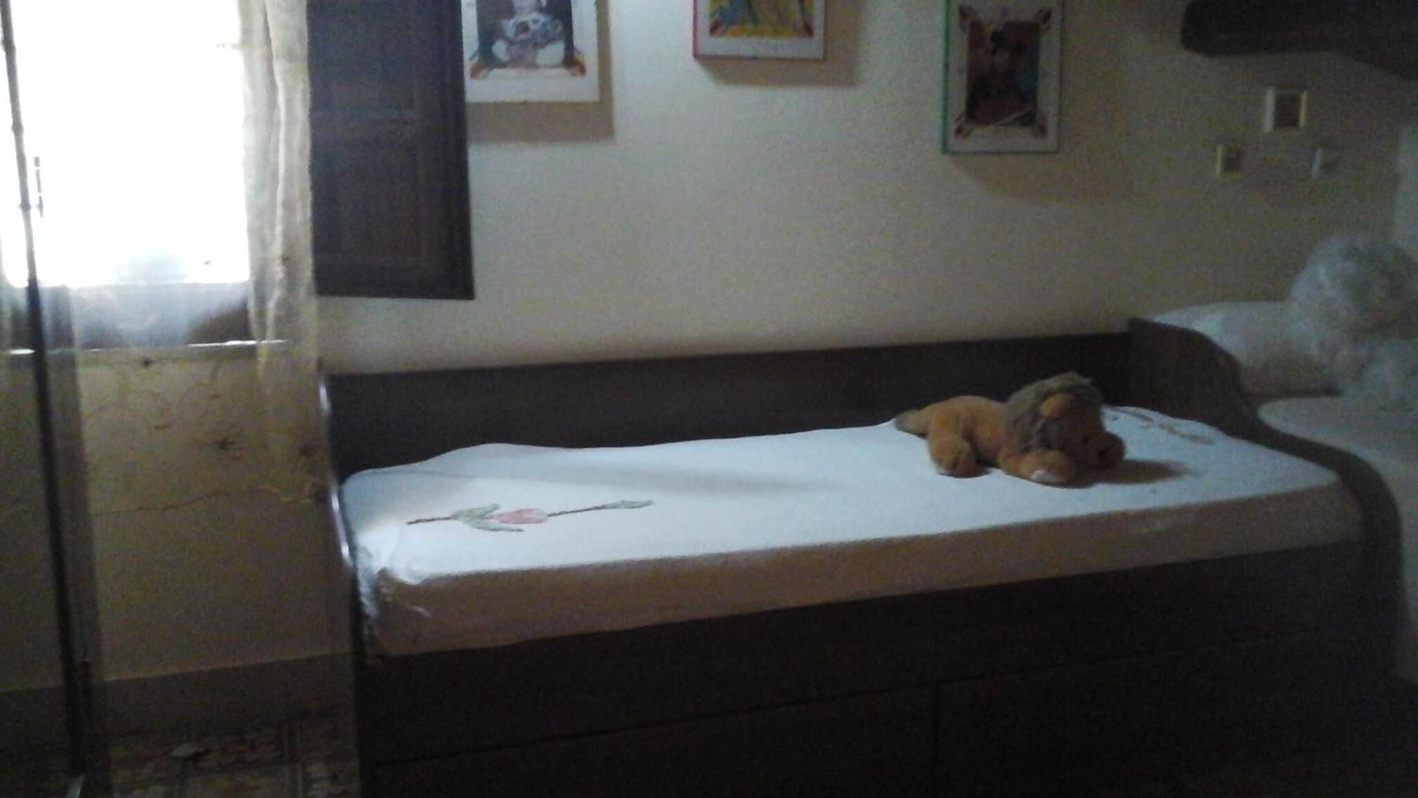 Una stanza piccola e scarsamente illuminata, con un letto singolo, una finestra con tende e alcune foto incorniciate appese al muro.
