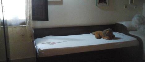 Una stanza piccola e scarsamente illuminata, con un letto singolo, una finestra con tende e alcune foto incorniciate appese al muro.
