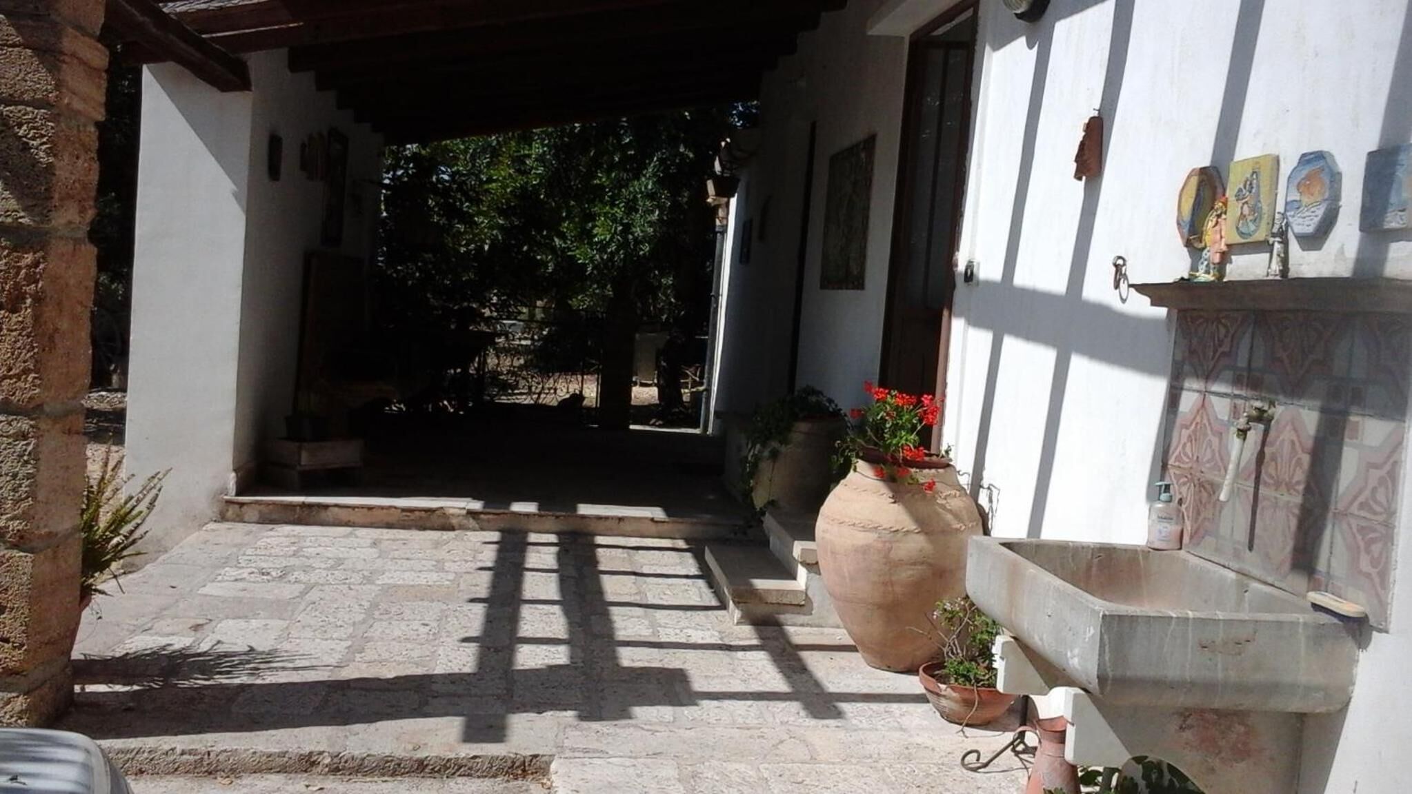 Une entrée dallée de pierres mène à un bâtiment doté d’un porche abrité, de plantes en pot et d’un mur recouvert de carreaux décorés d’œuvres d’art.