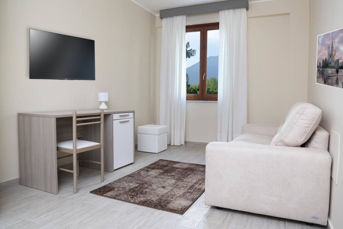 Una sala de estar moderna con un televisor de pantalla plana, un escritorio con una silla, un pequeño armario blanco, un sofá beige y vistas a montañas a través de una ventana con cortinas blancas.