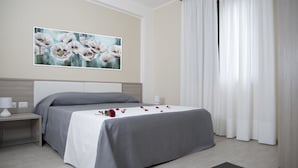 Habitación de hotel ordenada con cama, mesita de noche, lámpara, un cuadro de flores y un amplio ventanal con cortinas.