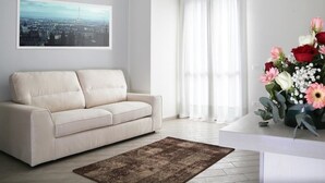 Una sala de estar moderna con un sofá color beige, una pintura de un paisaje urbano, un ventanal con cortinas traslúcidas y un arreglo floral sobre una mesa blanca.
