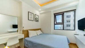 Un dormitorio con cama, escritorio, silla, televisor y ventana con vista a edificios.