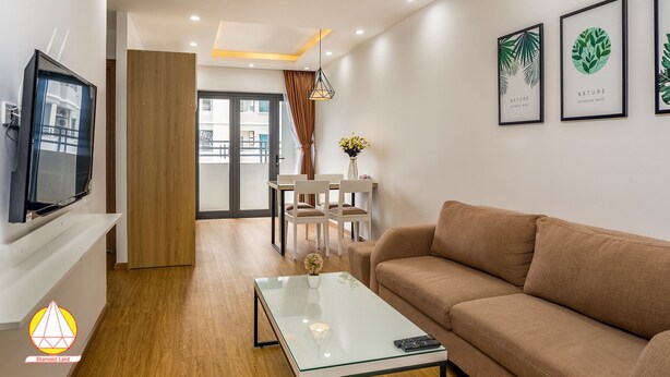 Una sala moderna con un sofá, una mesa de centro de vidrio y un televisor. Hay cuadros enmarcados en la pared y un comedor al fondo.