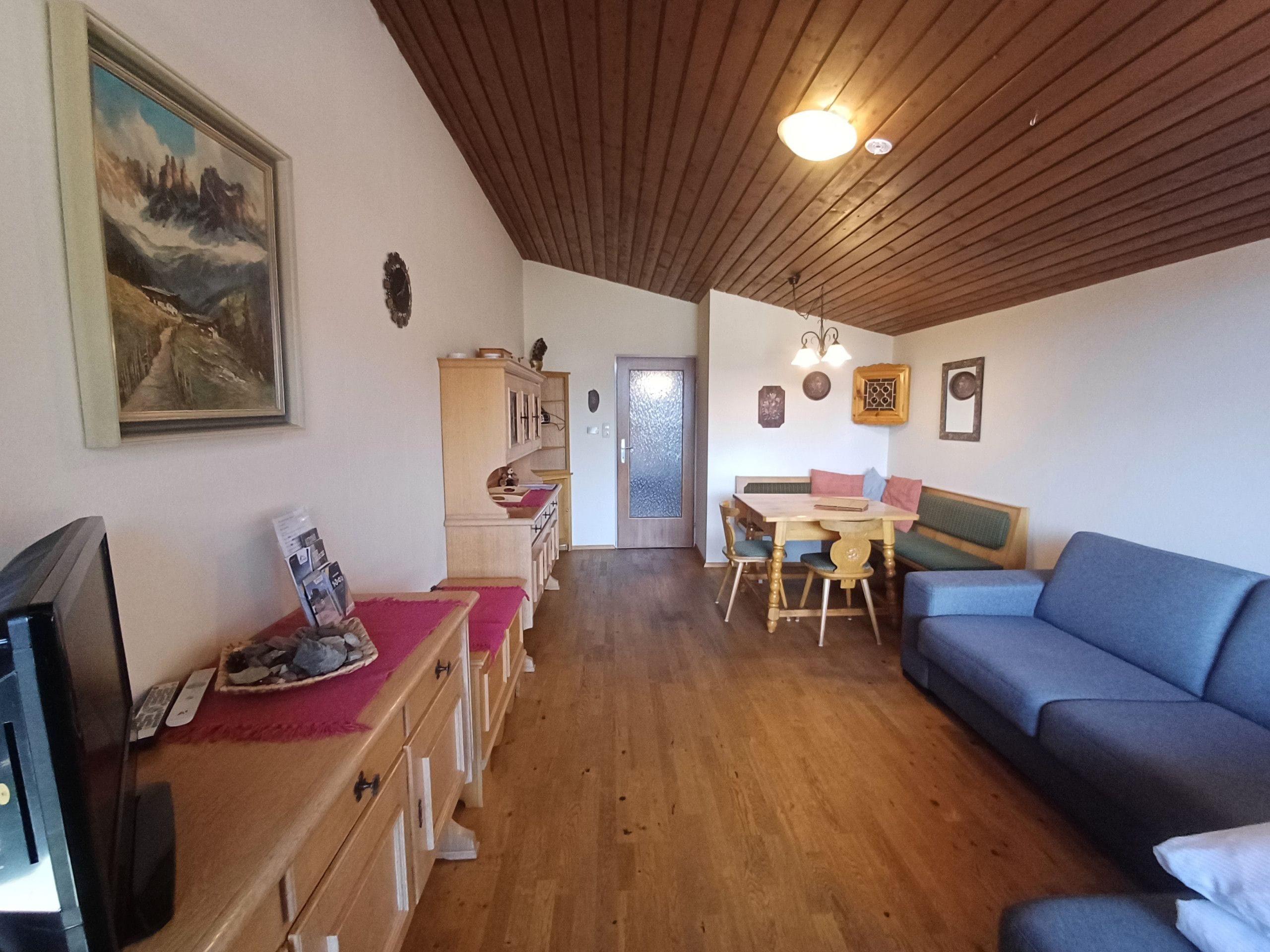 Un salón acogedor con techo de madera, un sofá azul, una mesa de comedor con sillas y un televisor a la izquierda.