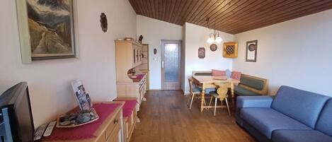 Un salón acogedor con techo de madera, un sofá azul, una mesa de comedor con sillas y un televisor a la izquierda.