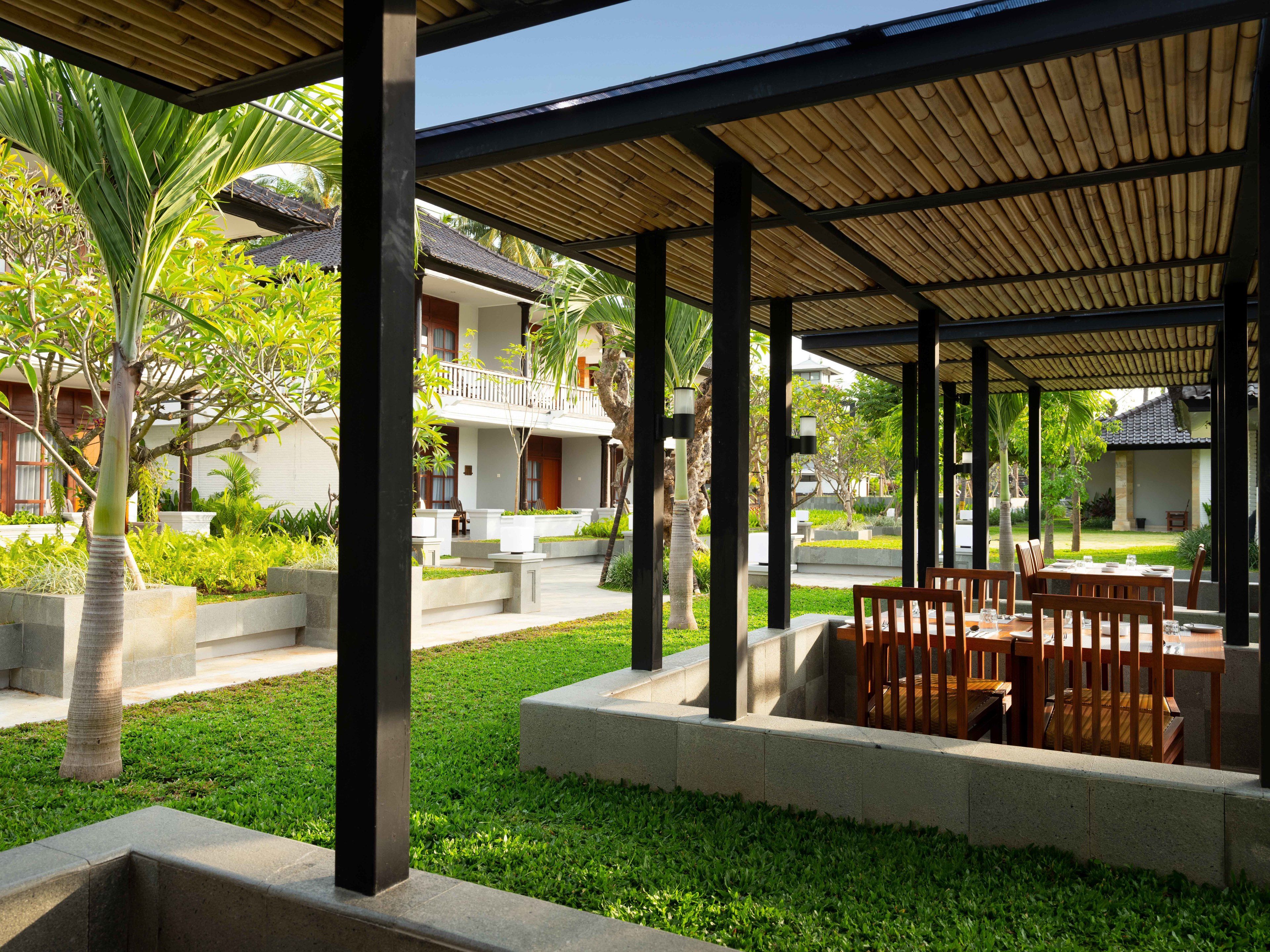 Un espace extérieur aménagé, abrité et doté de tables et de chaises, entouré de verdure et bordé d’un bâtiment avec un balcon.