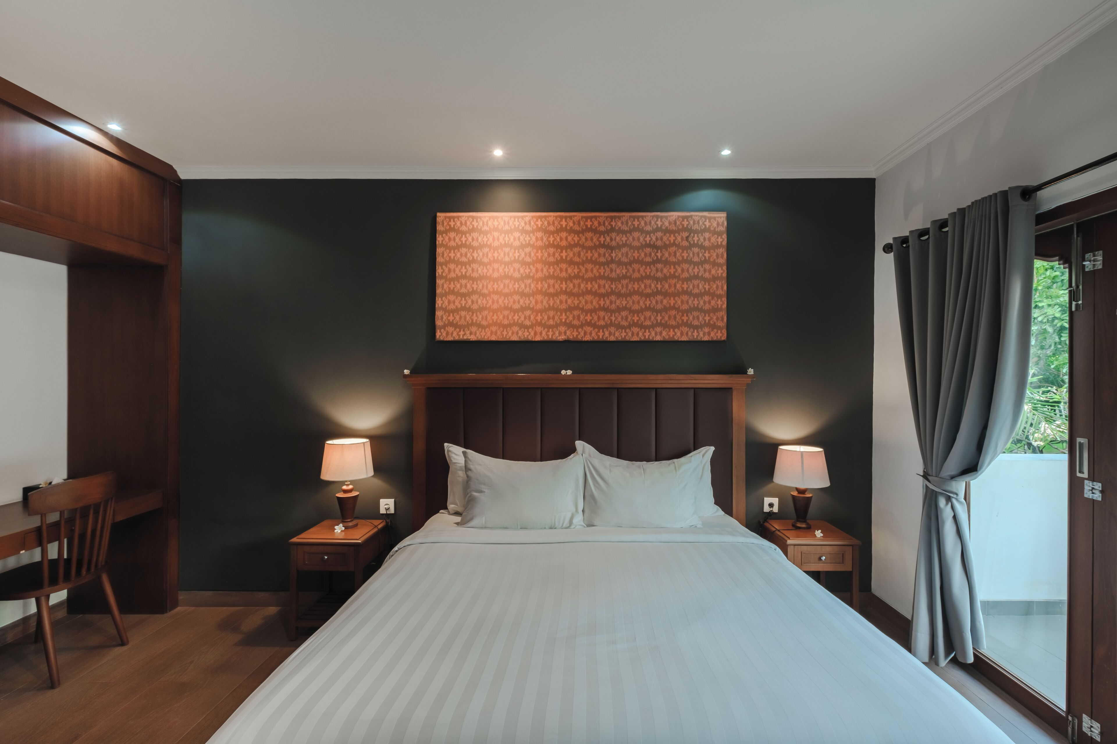 Habitación de hotel con una cama grande, dos mesitas de noche con lámparas, un cabecero de madera, una pared oscura y una ventana con cortinas.