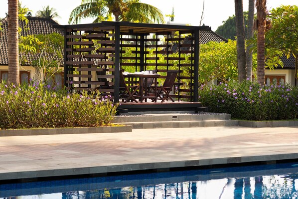 Zona de piscina con una pérgola de madera, una mesa con sillas y vistas a un edificio tradicional con tejado de tejas.