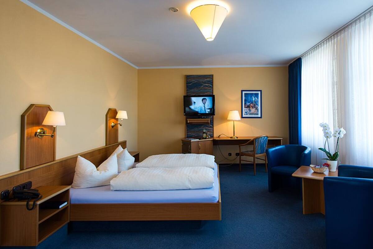A hotel room with a bed, bedside tables, a desk, a chair, a TV, and a window with curtains.