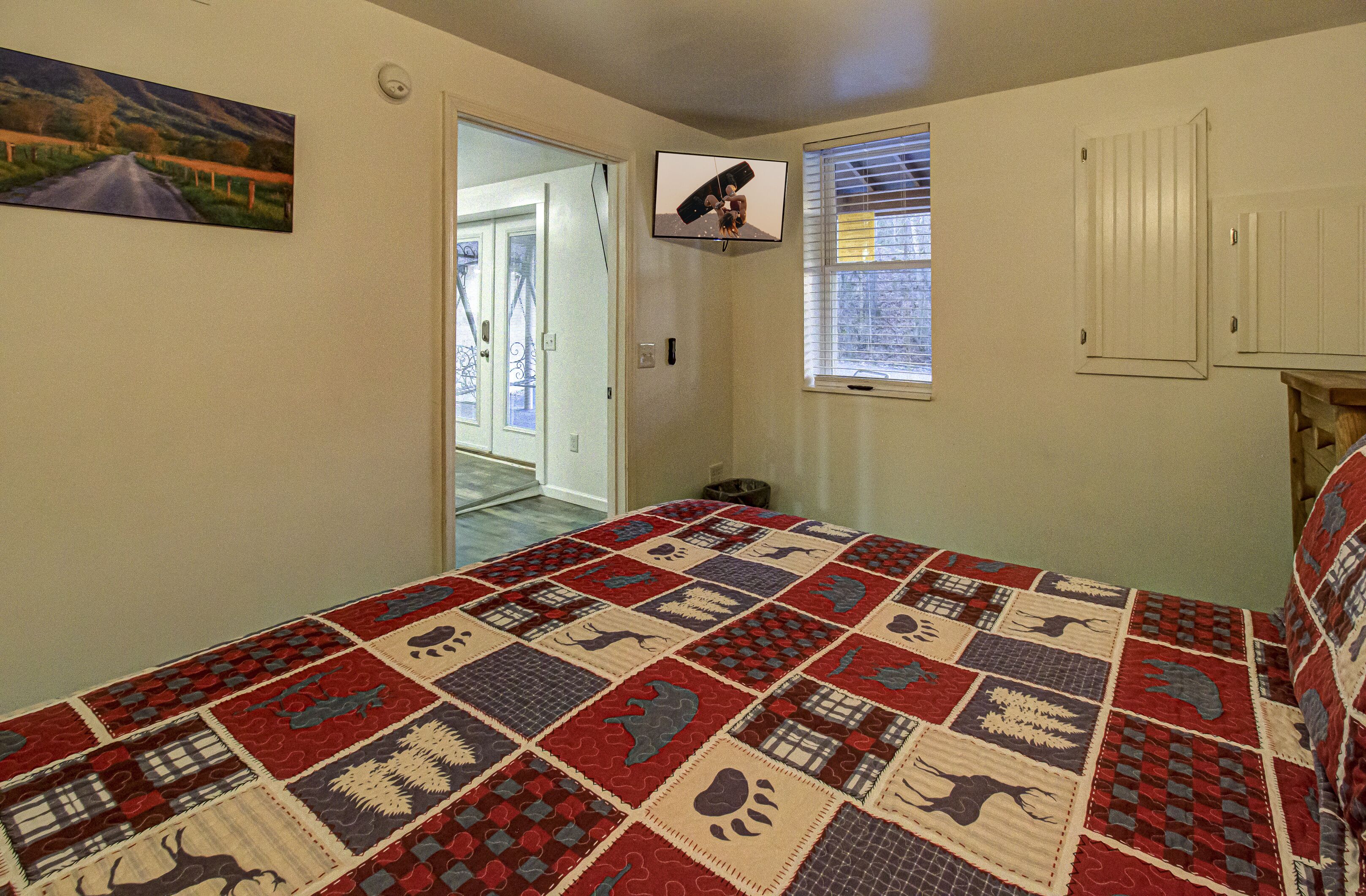 A bedroom with a large bed, a television mounted on the wall, a window with blinds, and a door leading to another room.