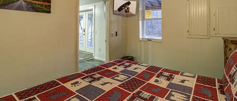 A bedroom with a large bed, a television mounted on the wall, a window with blinds, and a door leading to another room.
