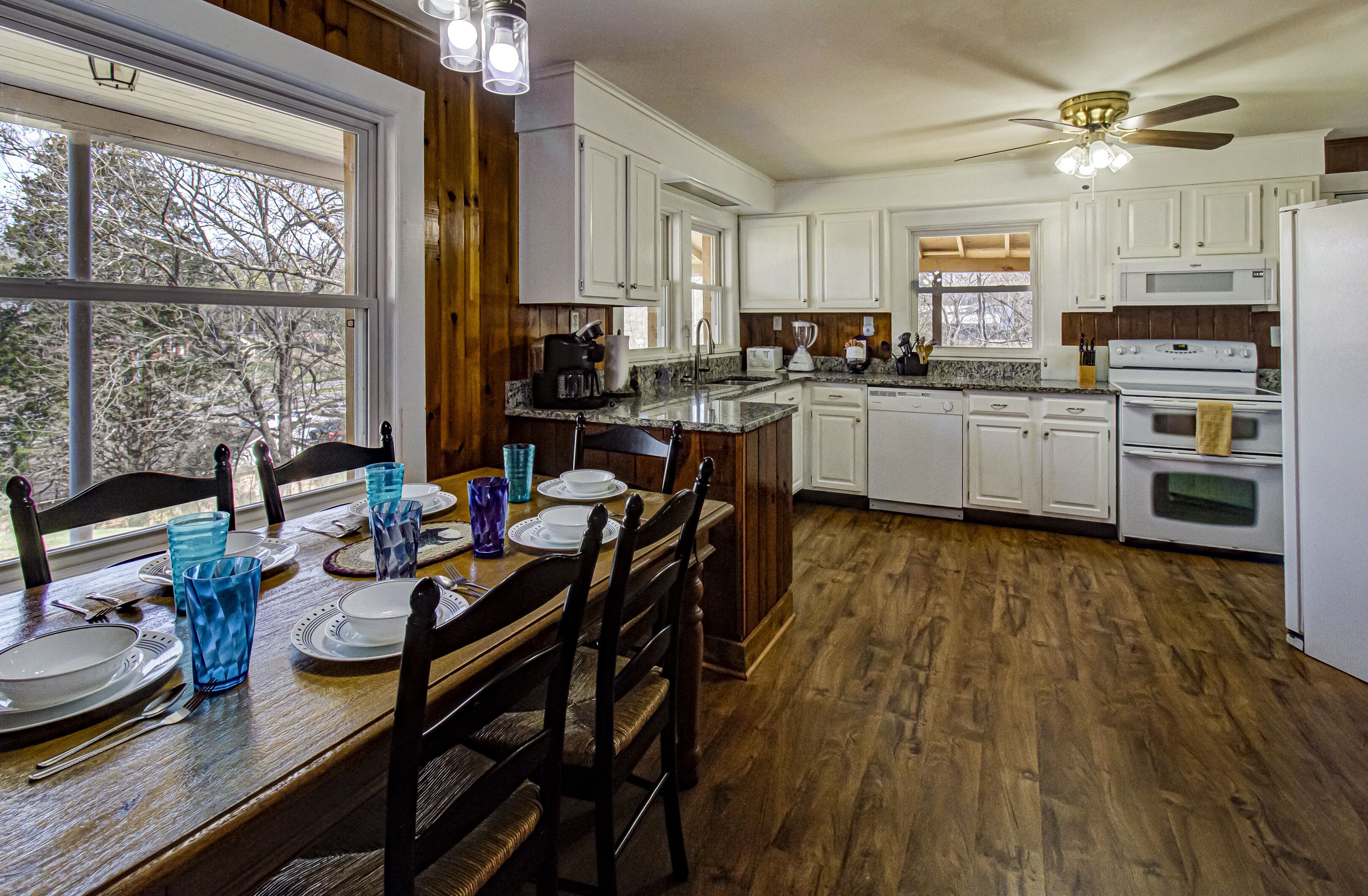 A kitchen with white cabinets, a wooden dining table set for four, and a large window.