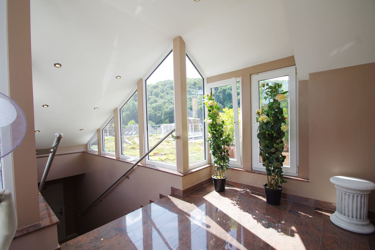 A spacious interior with a staircase leading to a balcony, large windows offering a view of greenery, and potted plants along the walls.